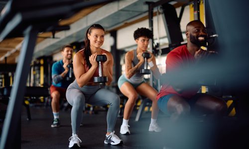 Happy multiracial athletes in squatting position exercising with dumbbells in health club. Focus is on young woman.